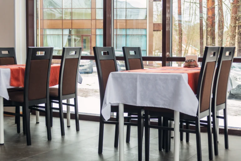 Salão de restaurante com mesas postas e cadeiras, sem pessoas, sob luz natural
