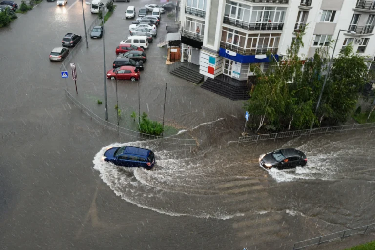 Rua de cidade do interior paulista alagada durante chuva forte, com carros atravessando a água no cruzamento.