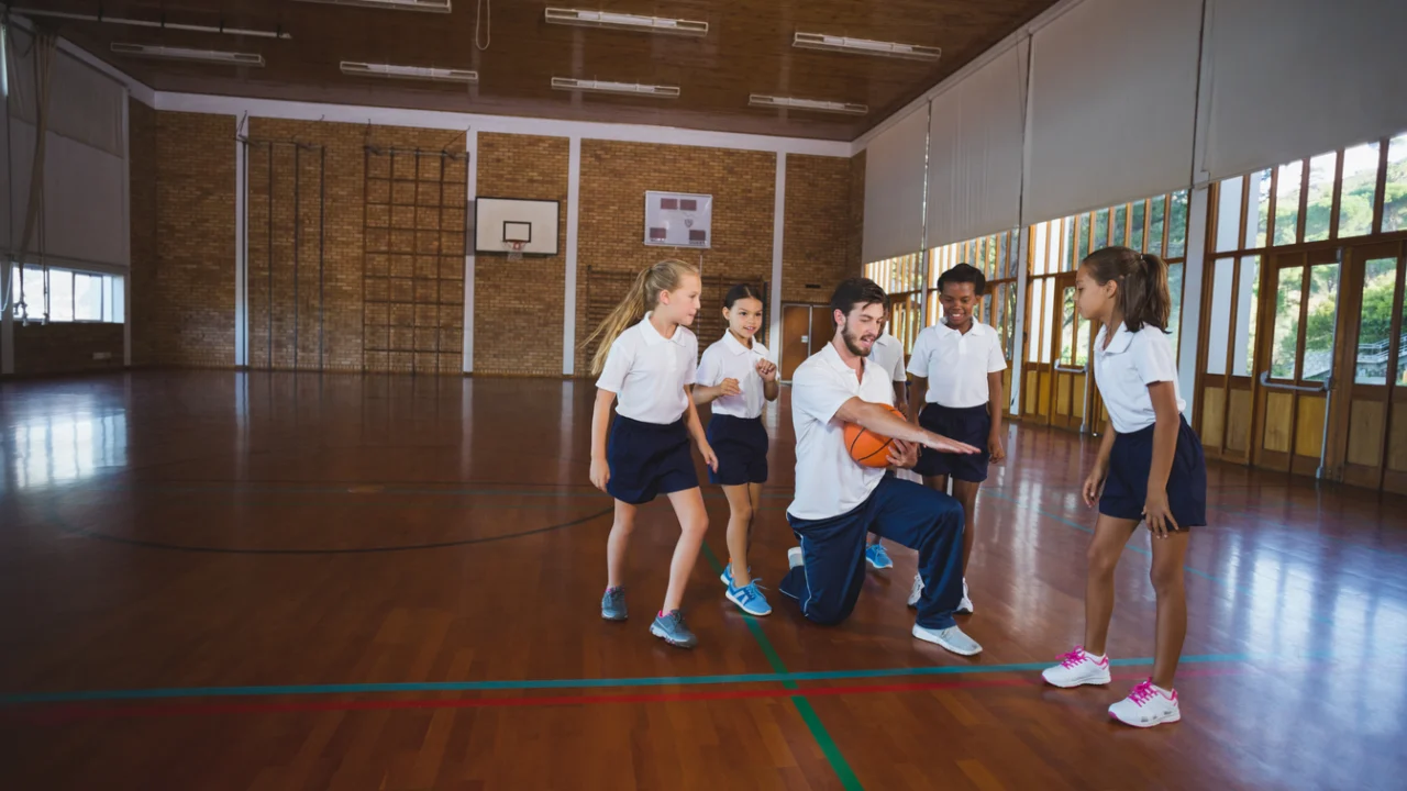 Professor de Educação Física com bola de basquete em quadra de escola, cercado por alunos uniformizados.