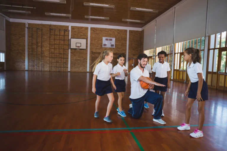 Professor de Educação Física com bola de basquete em quadra de escola, cercado por alunos uniformizados.