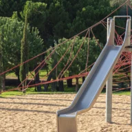 Escorregador metálico em pátio de recreação vazio, cercado por árvores, em manhã de sol