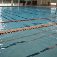 Piscina semiolímpica vazia com raias demarcadas em ginásio esportivo, com luz natural entrando pelas janelas