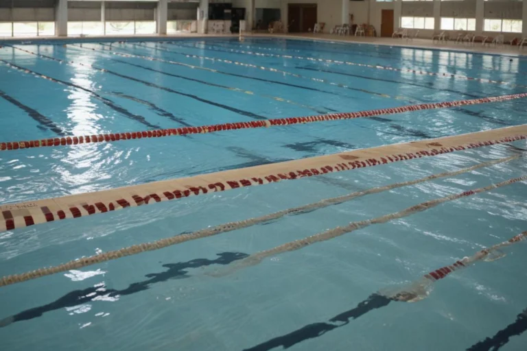 Piscina semiolímpica vazia com raias demarcadas em ginásio esportivo, com luz natural entrando pelas janelas