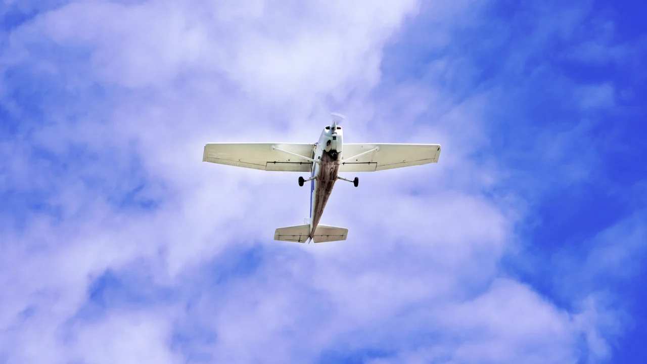 Avião monomotor branco em pleno voo contra um céu azul com nuvens