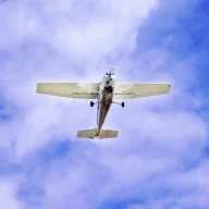 Avião monomotor branco em pleno voo contra um céu azul com nuvens