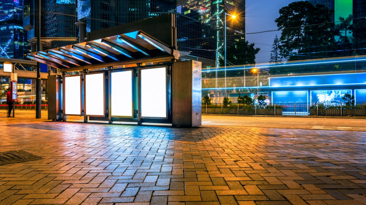 Calçada de terminal urbano à noite, com estrutura de vidro iluminada e prédios ao fundo