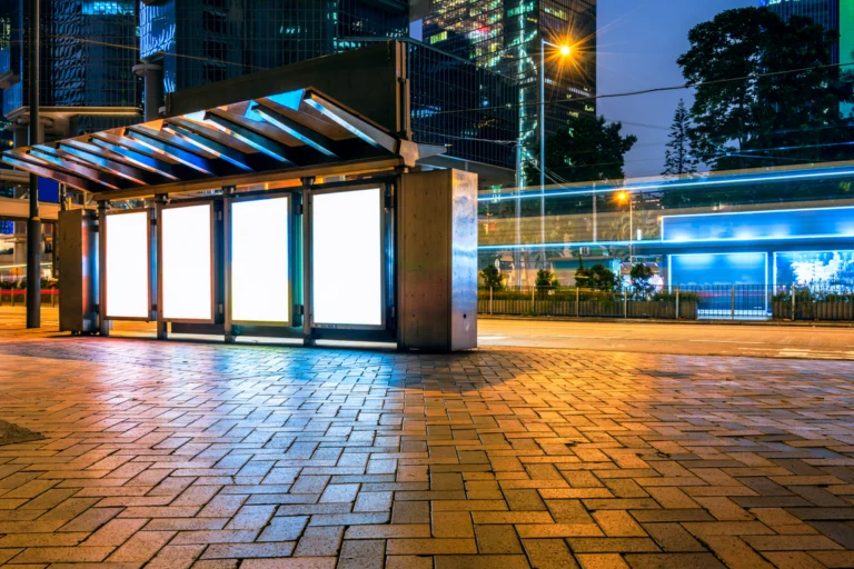 Calçada de terminal urbano à noite, com estrutura de vidro iluminada e prédios ao fundo