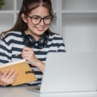 estudante sentada à mesa de biblioteca usando laptop, com cadernos abertos ao lado, luz natural da janela