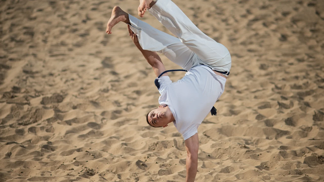 Atleta de capoeira executa movimento acrobático em área de areia