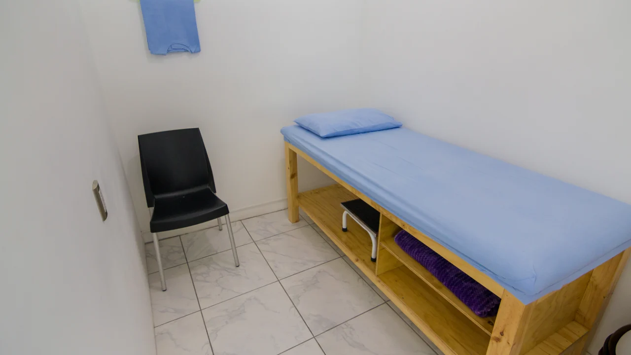 Sala de atendimento com maca coberta por lençol azul, cadeira e armário de madeira