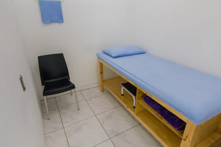 Sala de atendimento com maca coberta por lençol azul, cadeira e armário de madeira