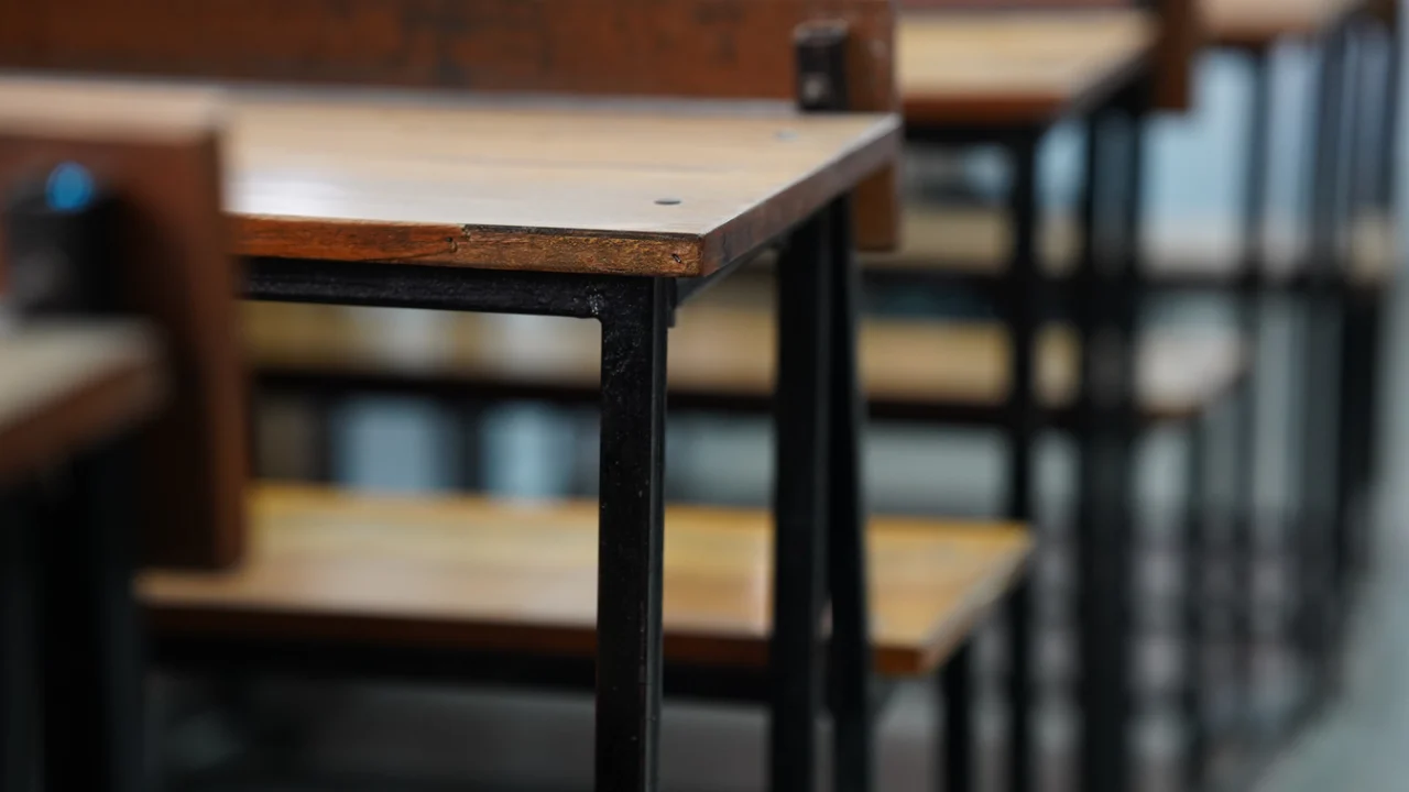 Carteiras escolares enfileiradas e vazias em uma sala de provas com canetas sobre as mesas