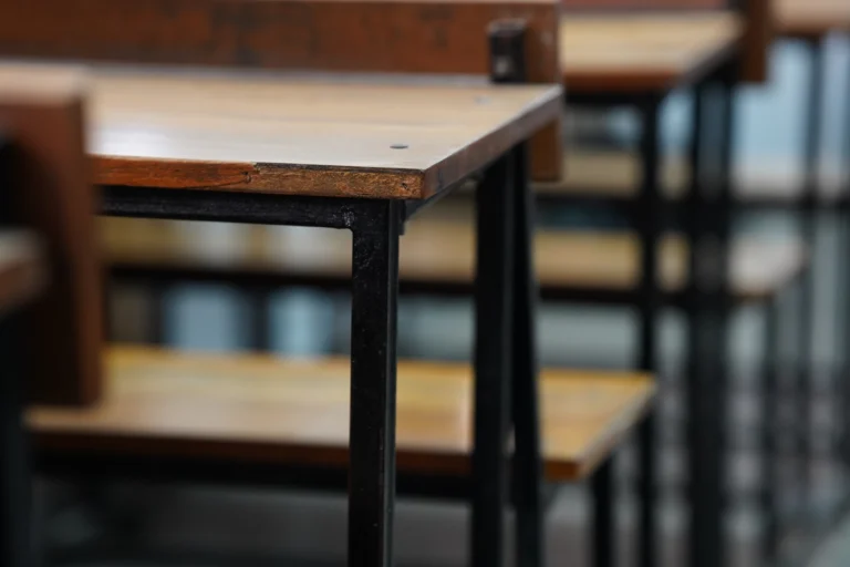 Carteiras escolares enfileiradas e vazias em uma sala de provas com canetas sobre as mesas