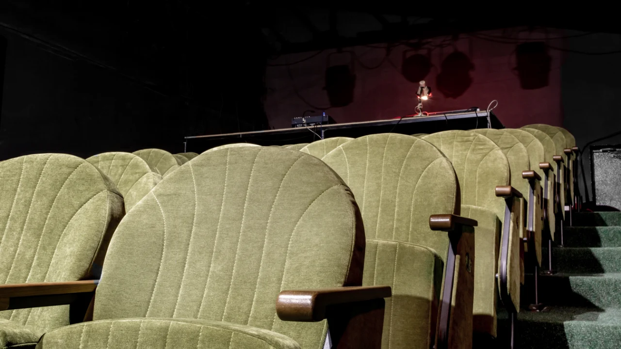 Poltronas verdes de teatro vazio, com palco iluminado ao fundo