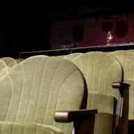 Poltronas verdes de teatro vazio, com palco iluminado ao fundo
