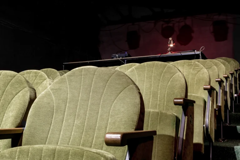 Poltronas verdes de teatro vazio, com palco iluminado ao fundo