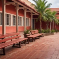 Pátio vazio de escola pública com muros vermelhos, bancos de madeira, piso de tijolos e palmeiras em manhã de sol