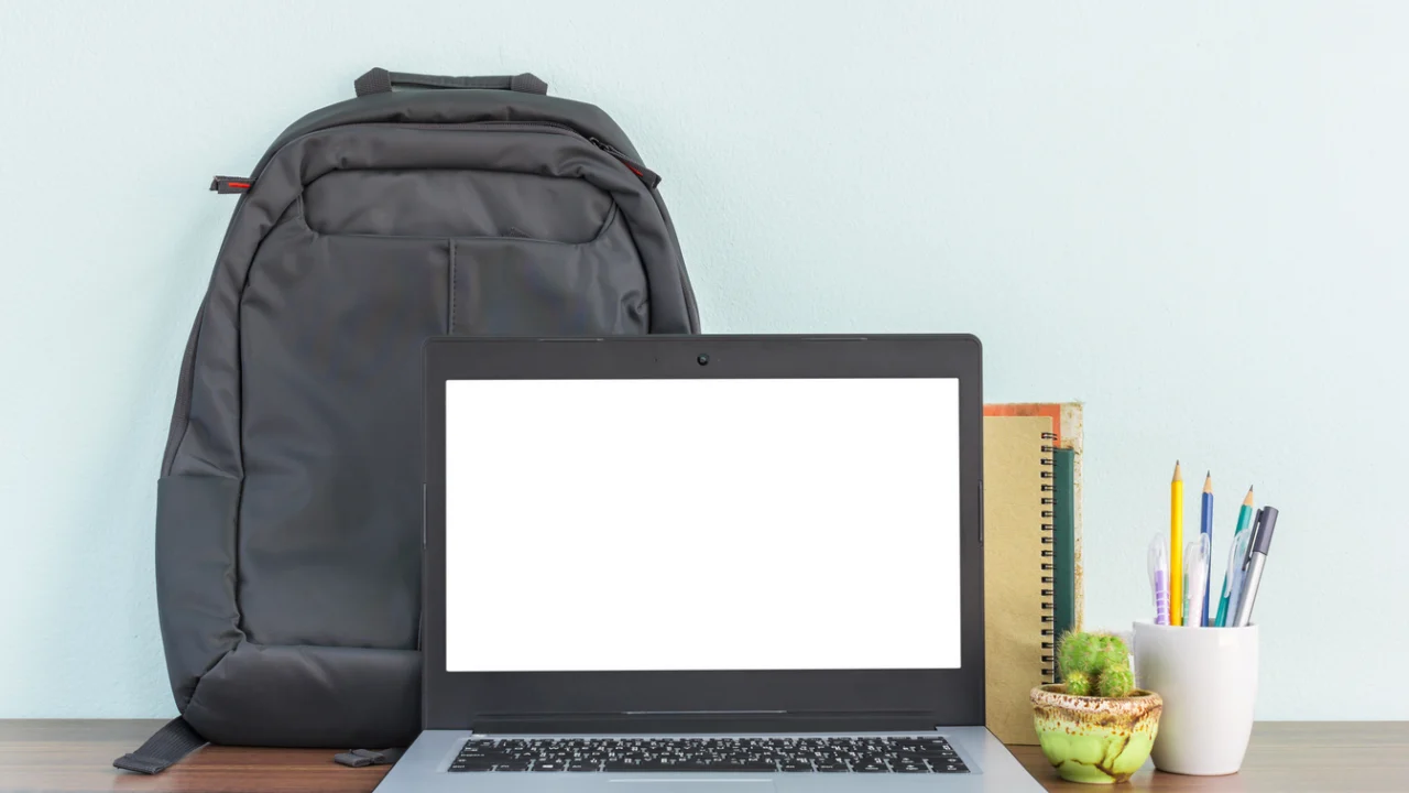 Mesa de estudos com notebook aberto, mochila ao fundo e cadernos e canetas ao lado