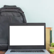 Mesa de estudos com notebook aberto, mochila ao fundo e cadernos e canetas ao lado