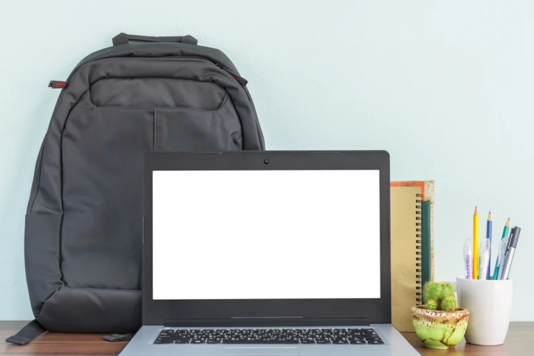 Mesa de estudos com notebook aberto, mochila ao fundo e cadernos e canetas ao lado