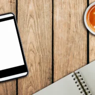 celular apoiado sobre mesa de madeira com xícara de café e caderno, em luz natural
