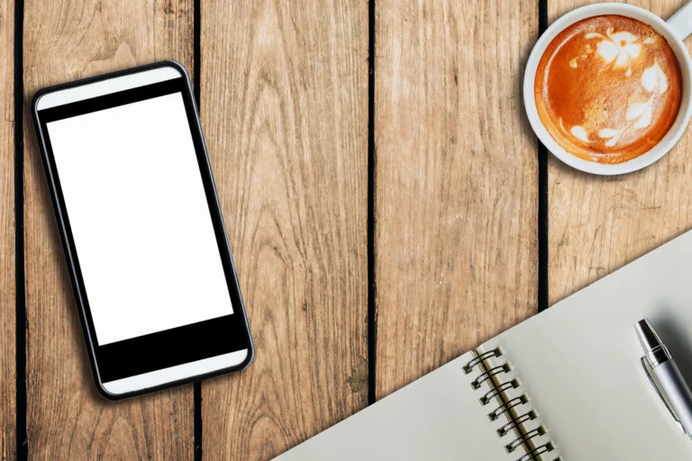 celular apoiado sobre mesa de madeira com xícara de café e caderno, em luz natural