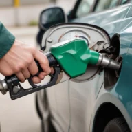 Mão segurando o bico de abastecimento verde ao lado da tampa do tanque de um carro em um posto de combustível, durante o dia