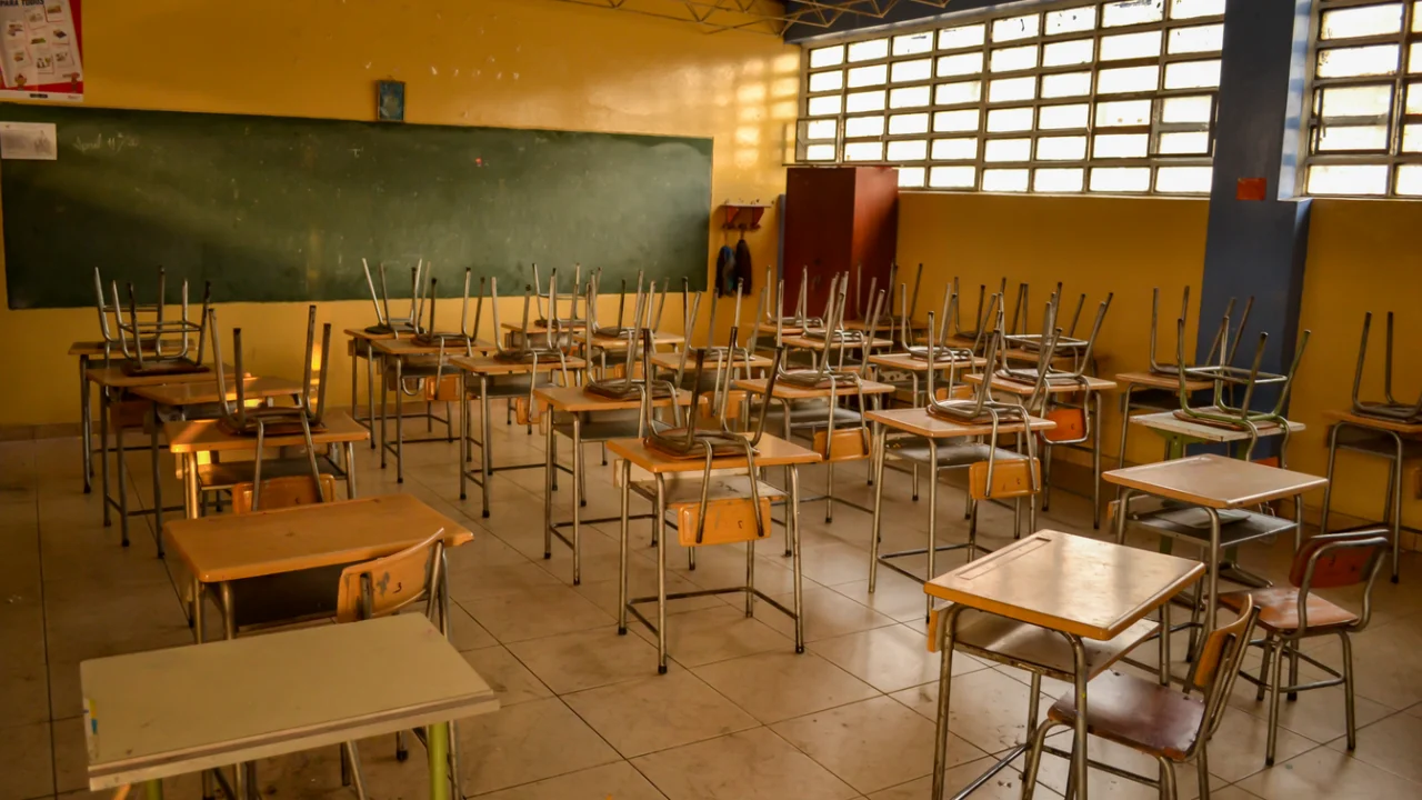 Carteiras enfileiradas em sala de aula de escola pública, com quadro ao fundo
