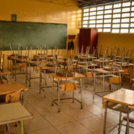 Carteiras enfileiradas em sala de aula de escola pública, com quadro ao fundo
