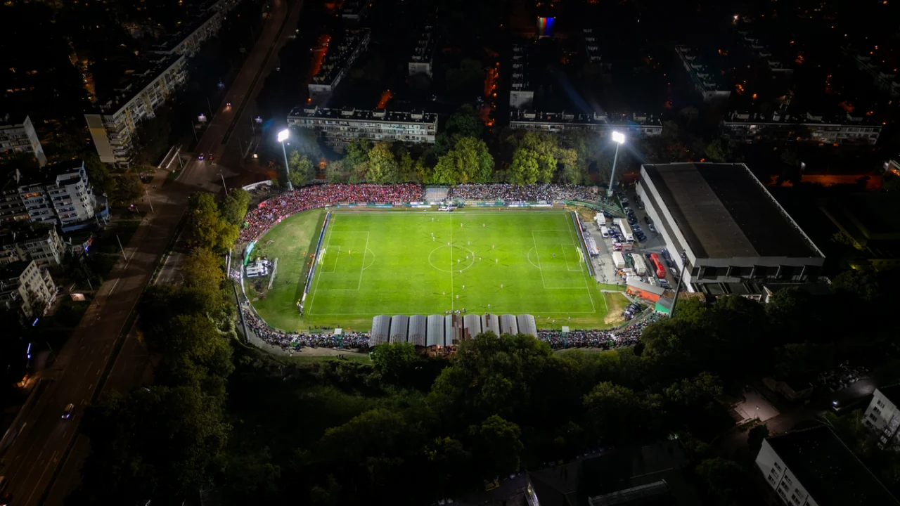 Estádio de futebol iluminado à noite, visto de cima, com o gramado verde e a bola parada no centro do campo