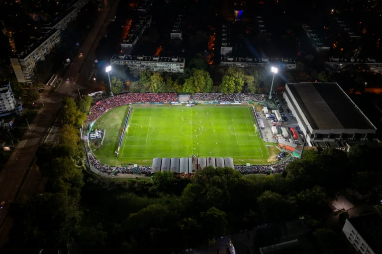 Estádio de futebol iluminado à noite, visto de cima, com o gramado verde e a bola parada no centro do campo
