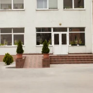 Fachada de prédio escolar com janelas amplas, entrada com escada e pequenos arbustos em vasos