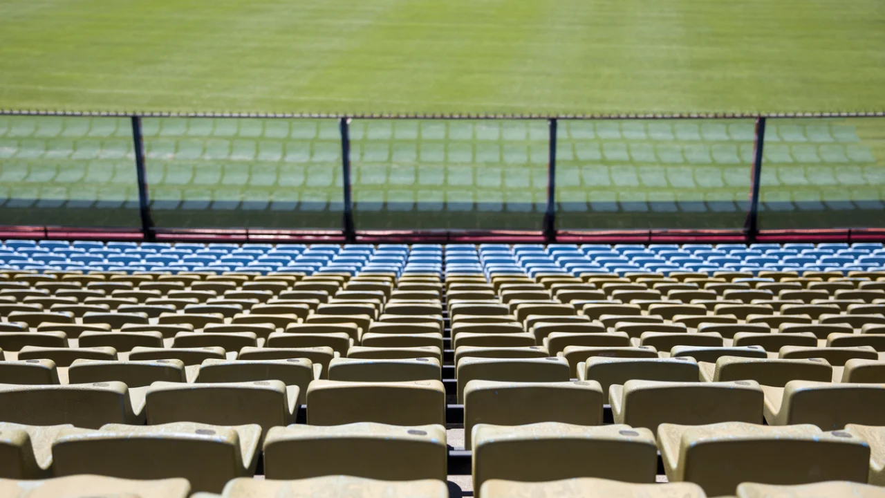 Arquibancadas vazias de estádio de futebol com cadeiras amarelas e azuis e o gramado ao fundo