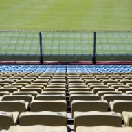 Arquibancadas vazias de estádio de futebol com cadeiras amarelas e azuis e o gramado ao fundo