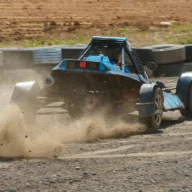 Carro de competição em pista de terra levantando poeira durante uma prova, com barreira de pneus ao fundo