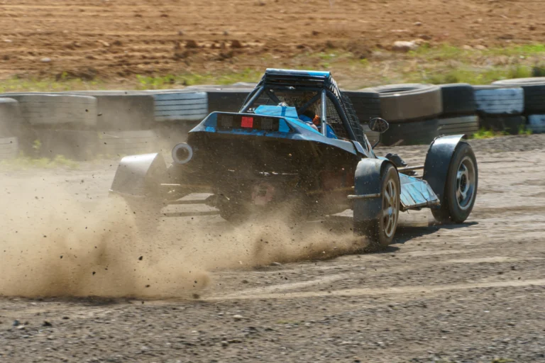 Carro de competição em pista de terra levantando poeira durante uma prova, com barreira de pneus ao fundo