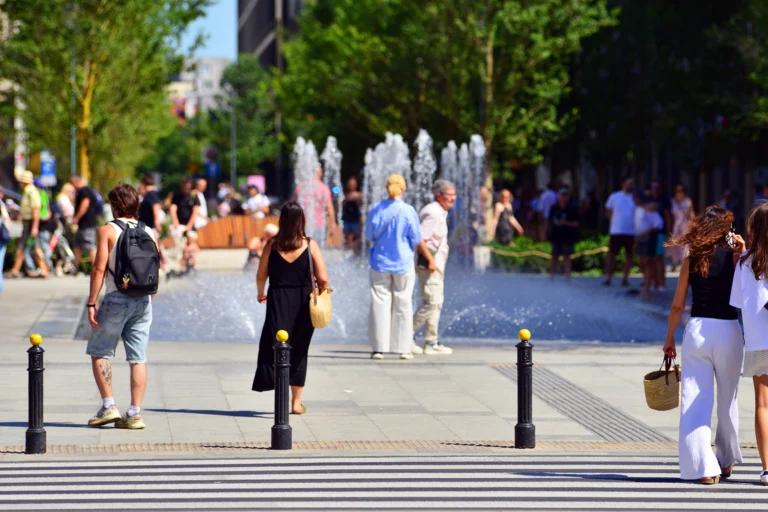Pessoas caminhando em uma praça central arborizada com fonte, em manhã de sol