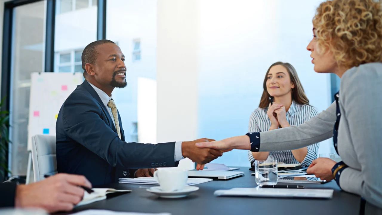 Duas pessoas apertando as mãos sobre uma mesa de reunião em sala clara