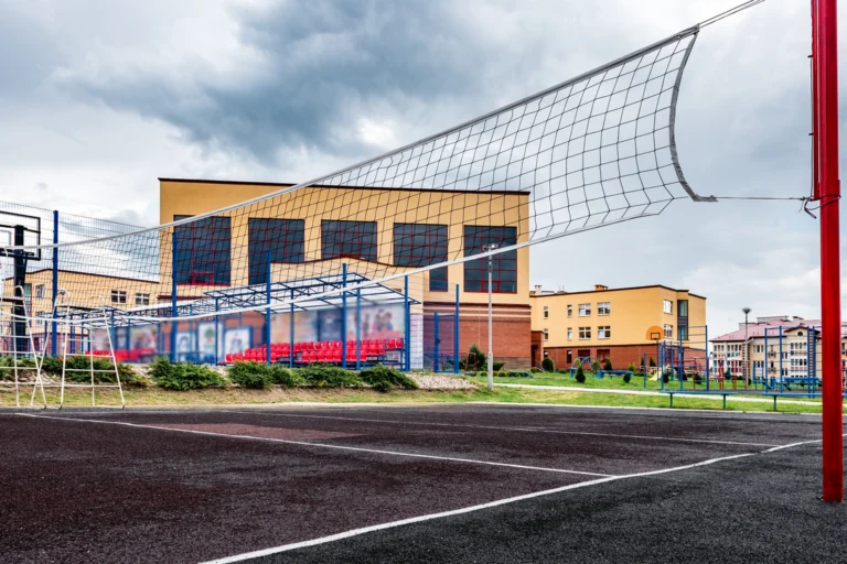 Quadra esportiva externa de escola pública, com rede e cesta de basquete, ao lado do prédio escolar