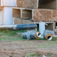 Tubos de concreto e valas abertas em canteiro de obras, com área verde e prédio em construção ao fundo