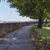 Calçadão arborizado às margens de rio, com mureta de concreto e bancos