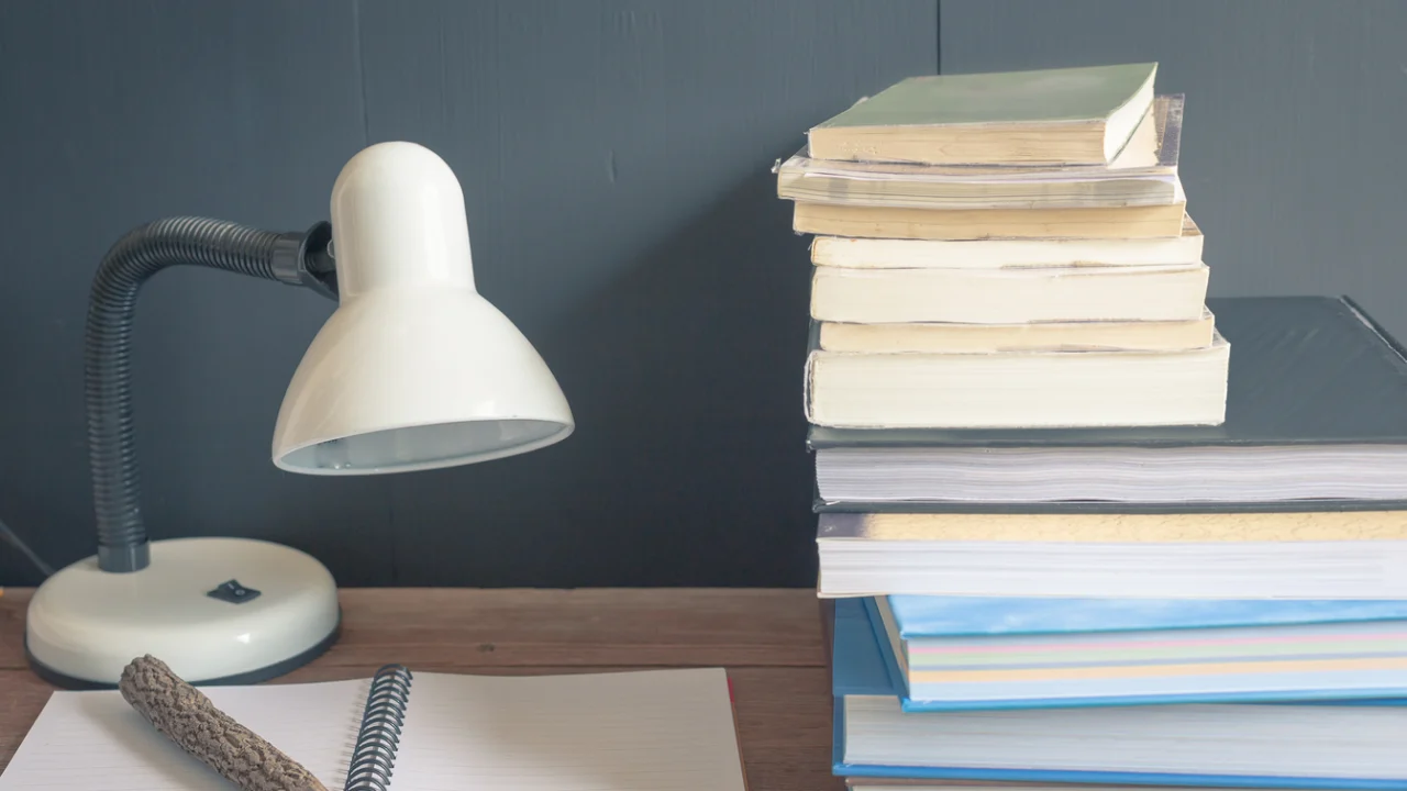 Livros, caderno e luminária sobre uma mesa de estudos.
