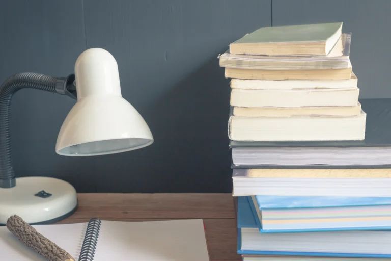 Livros, caderno e luminária sobre uma mesa de estudos.