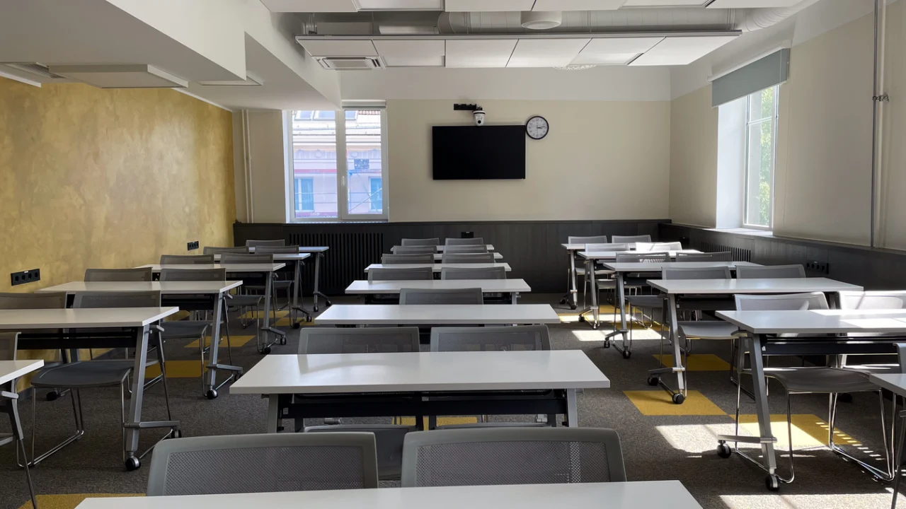Sala de aula vazia com mesas e cadeiras iluminadas pelas janelas.