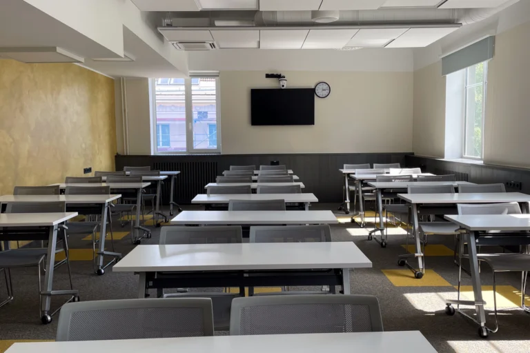 Sala de aula vazia com mesas e cadeiras iluminadas pelas janelas.