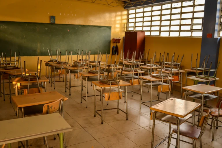 Sala de aula de escola pública com carteiras e cadeiras enfileiradas vazias, iluminada pela luz da manhã