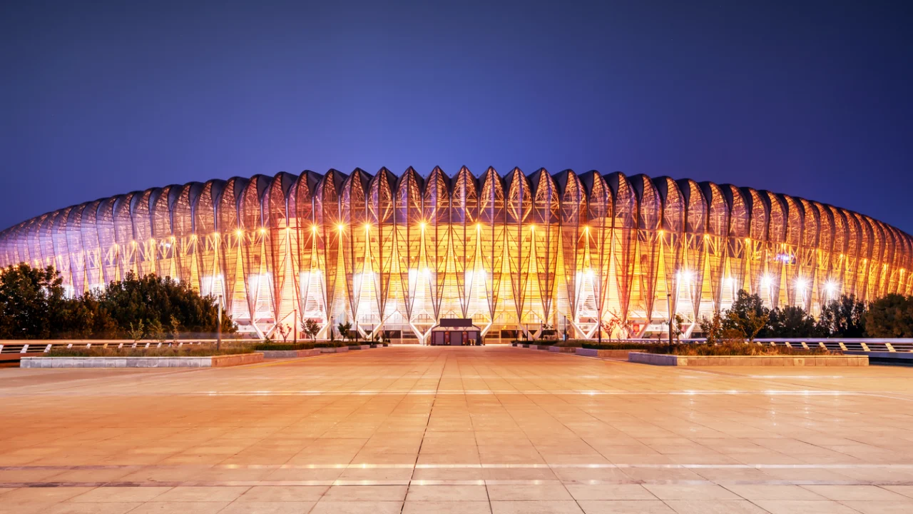 Exterior de um grande estádio urbano iluminado ao anoitecer, com praça de acesso vazia