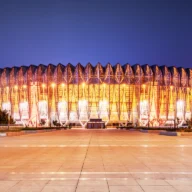Exterior de um grande estádio urbano iluminado ao anoitecer, com praça de acesso vazia