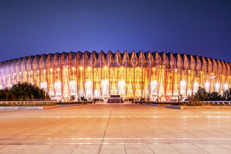 Exterior de um grande estádio urbano iluminado ao anoitecer, com praça de acesso vazia