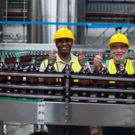 Dois trabalhadores de capacete e colete refletivo em linha de produção de bebidas em uma fábrica